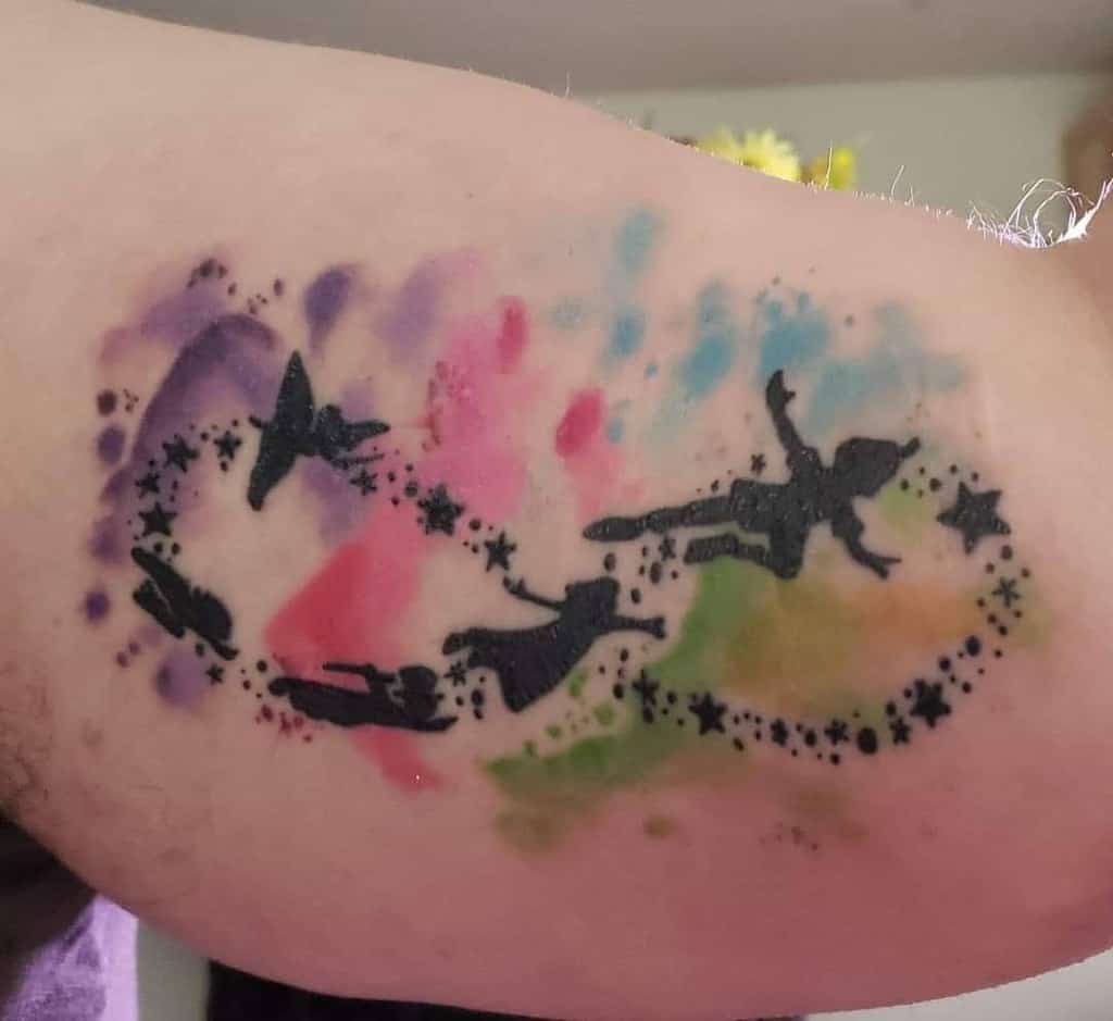 A whimsical watercolor-inspired tattoo of silhouetted figures flying in a starry arc, set against a vibrant splash of colors