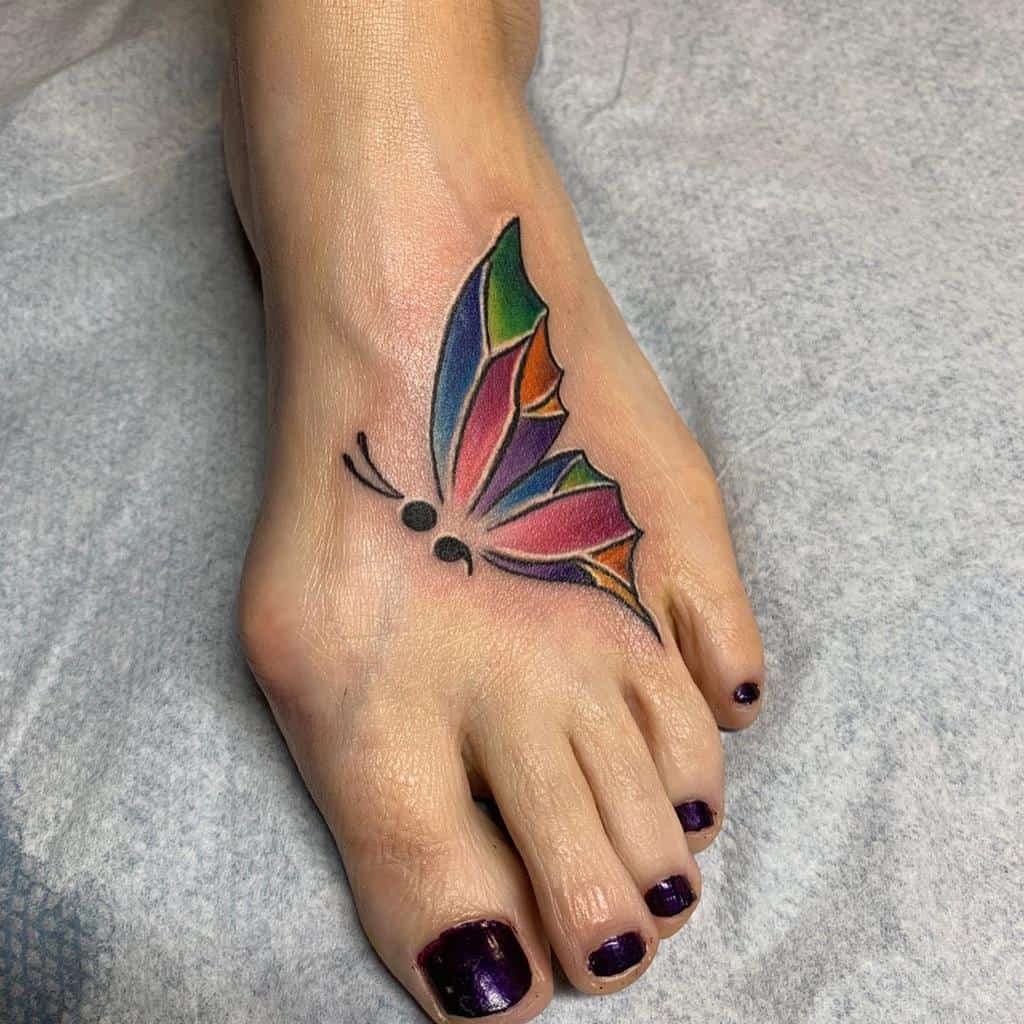 A colorful semicolon butterfly tattoo on the foot, featuring bright shades of orange, pink, and green, symbolizing personal growth, mental health awareness, and the beauty of transformation