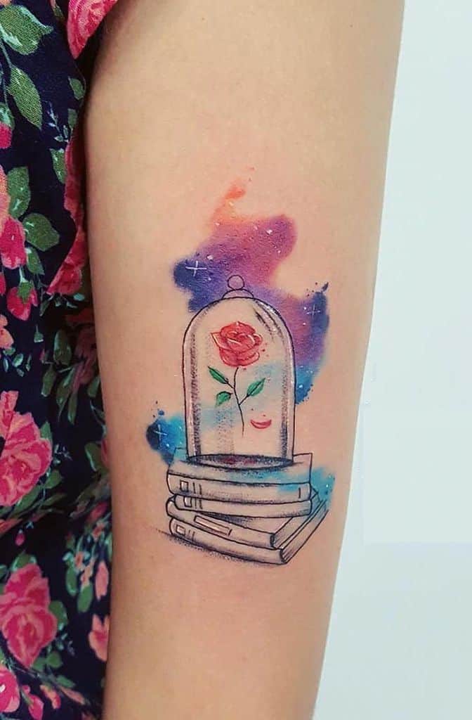 Watercolor-style tattoo of the enchanted rose inside a glass dome, placed on a stack of books, with vibrant splashes of blue, purple, and orange.