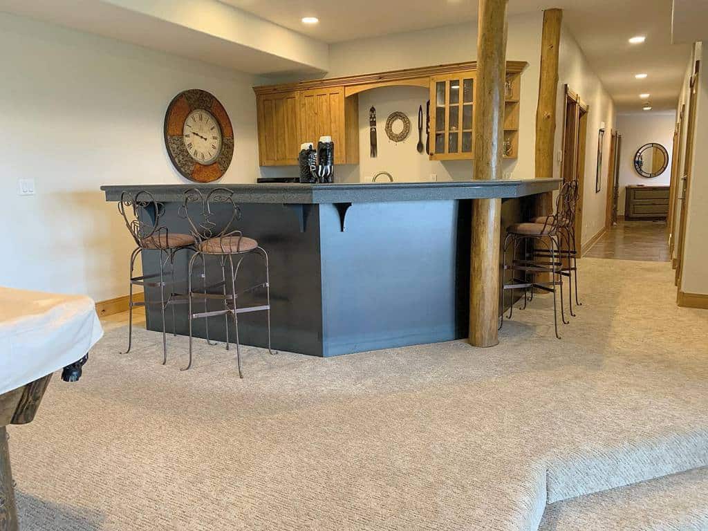 Rustic corner basement bar with wooden posts, metal stools, and cozy neutral carpeted flooring.