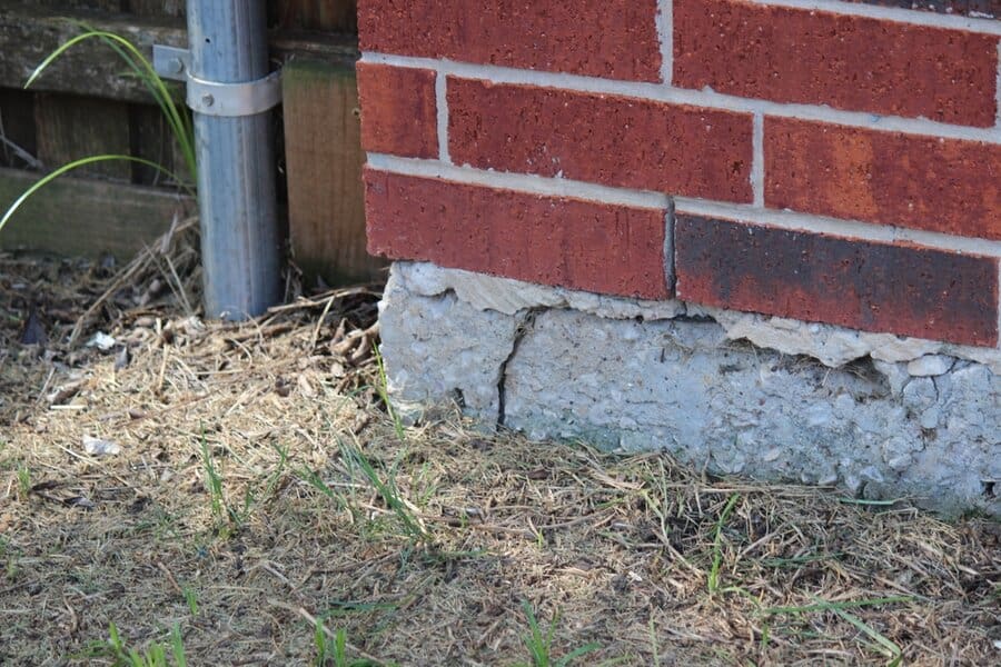 Close-up of a house foundation with a visible horizontal crack below a red brick wall, next to dry grass and soil