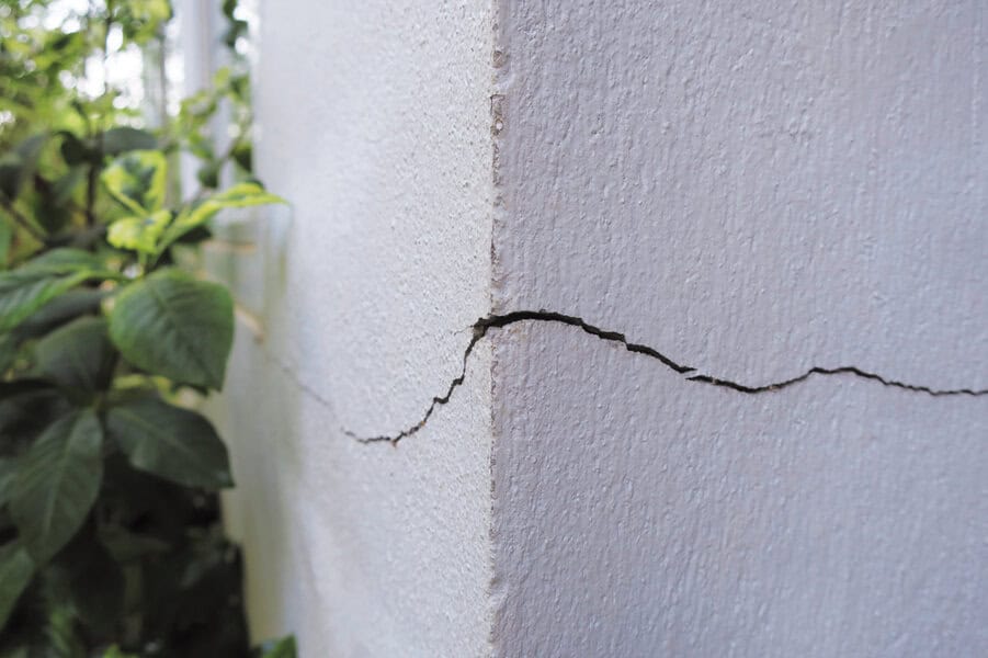 A large crack runs along the corner of a white exterior wall, with green plants visible on the left side