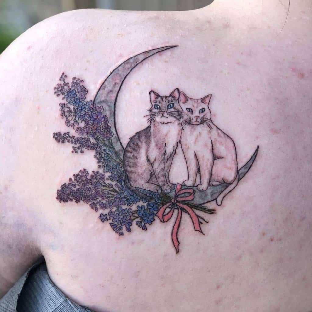 Tattoo of two cats sitting on a crescent moon adorned with flowers and a pink ribbon