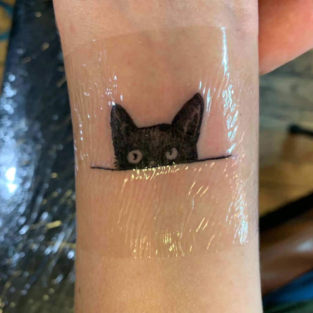 A tattoo of a black cat peeking over the edge, captures its adorable curiosity