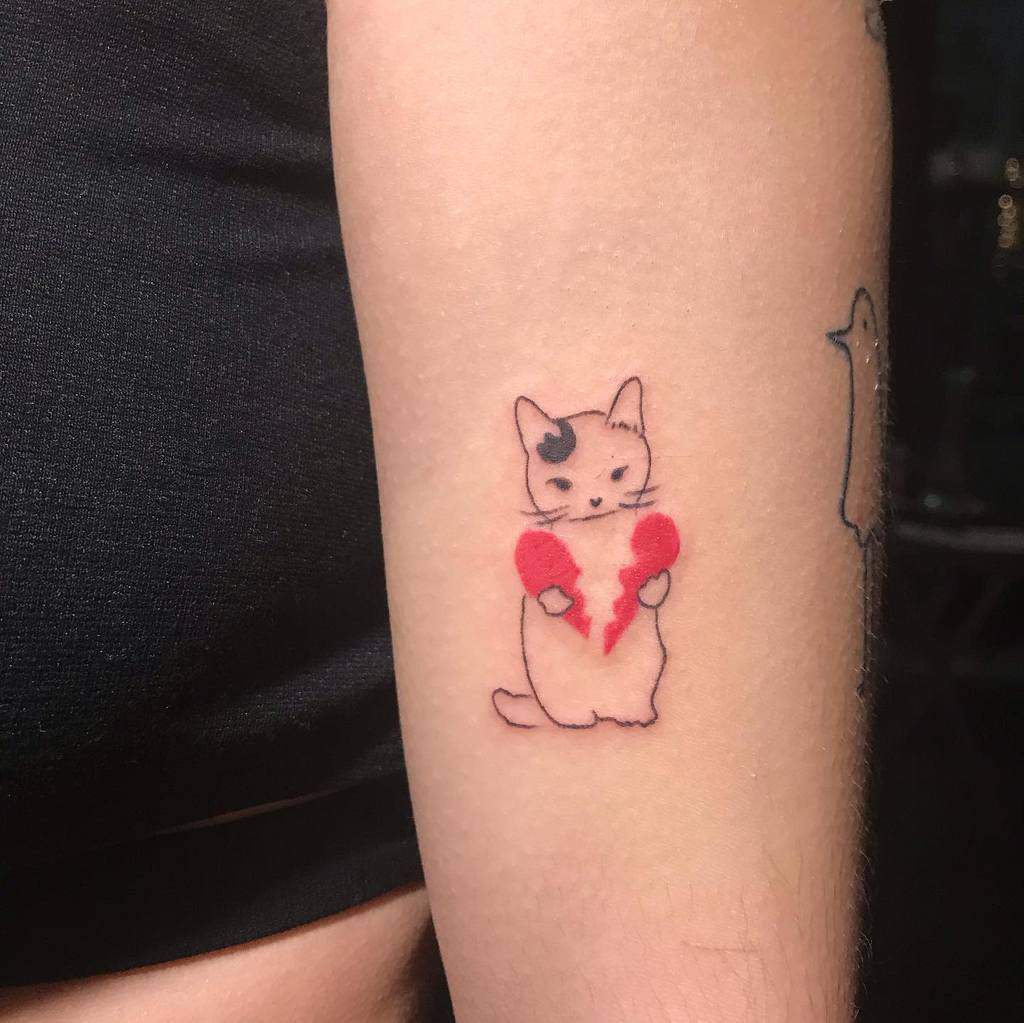 Tattoo of a playful cat holding a broken red heart on a person's arm