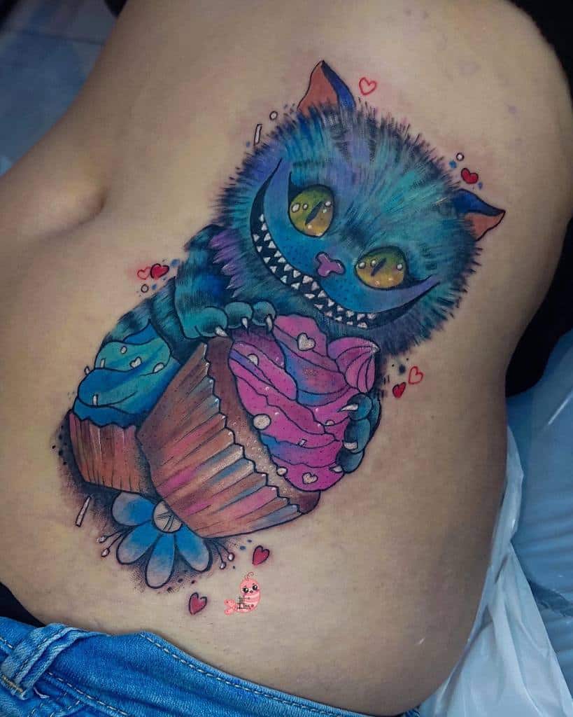 A colorful and playful Cheshire Cat tattoo holding two cupcakes, surrounded by small hearts and details
