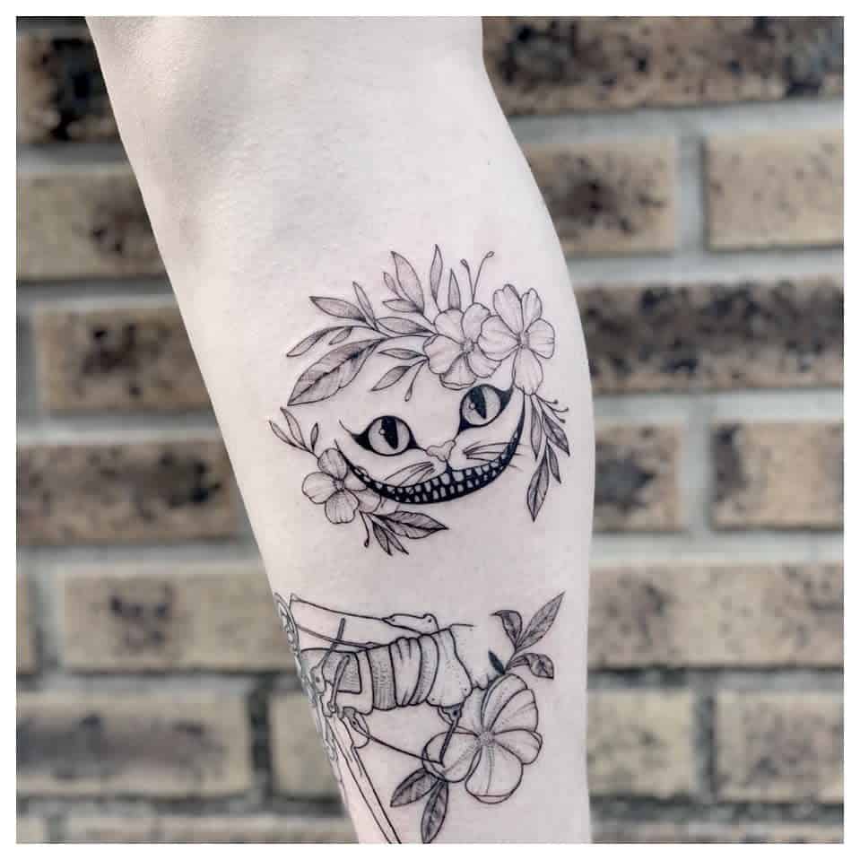 A delicate black ink Cheshire Cat tattoo, featuring its iconic grin and eyes adorned with floral details