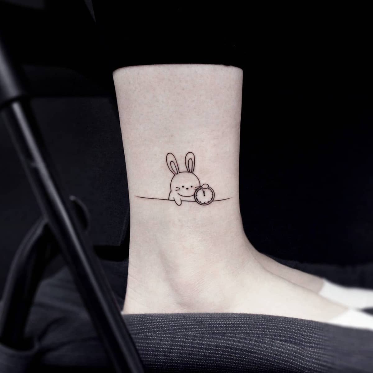 Simple tattoo of a cute bunny holding a pocket watch on an ankle
