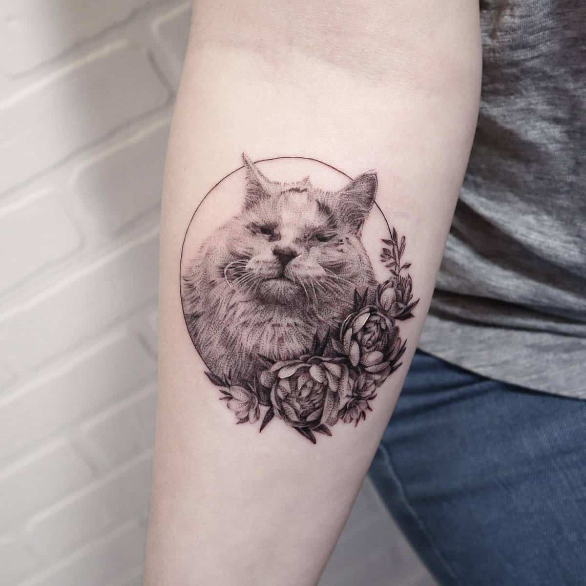 Forearm tattoo of a fluffy cat with flowers around it, done in black and gray ink