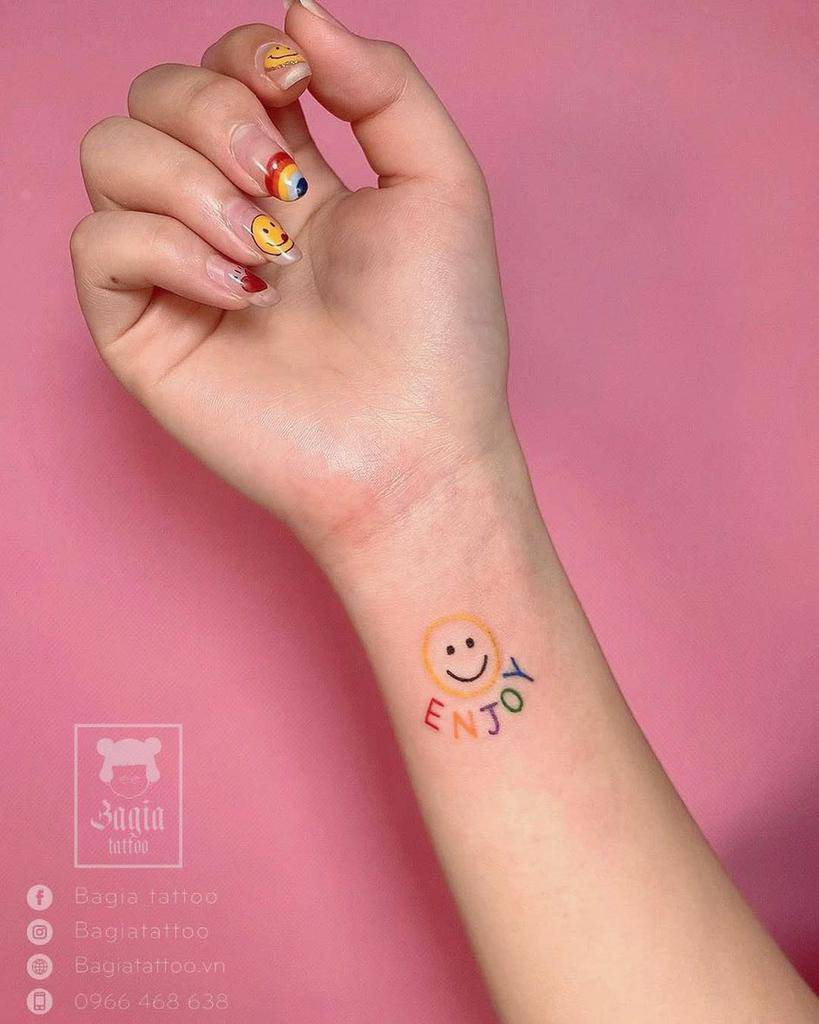A wrist tattoo with a smiley face and "ENJOY" in colorful letters