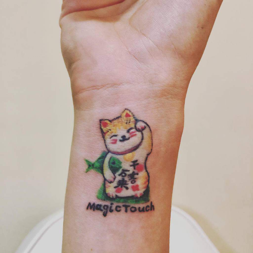 Cute maneki-neko tattoo on a wrist with the words "Magic Touch" below it