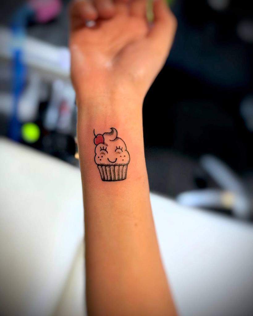 Cute cupcake tattoo with a smiley face and cherry on top, inked on a wrist