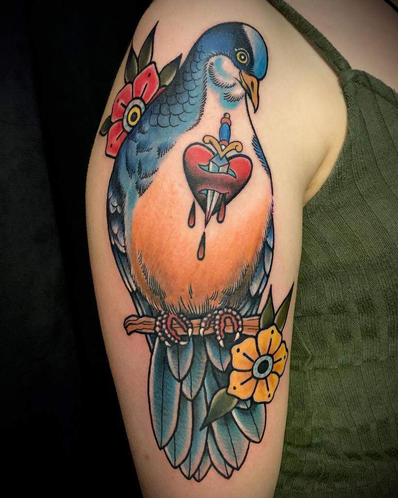 Tattoo of a blue bird on a branch with a bleeding heart pierced by a dagger and surrounded by flowers