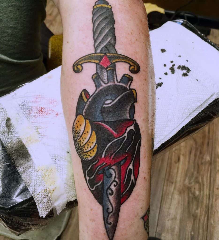 Tattoo of a dagger piercing a bleeding heart with a steel blade and ornate handle on a person's forearm