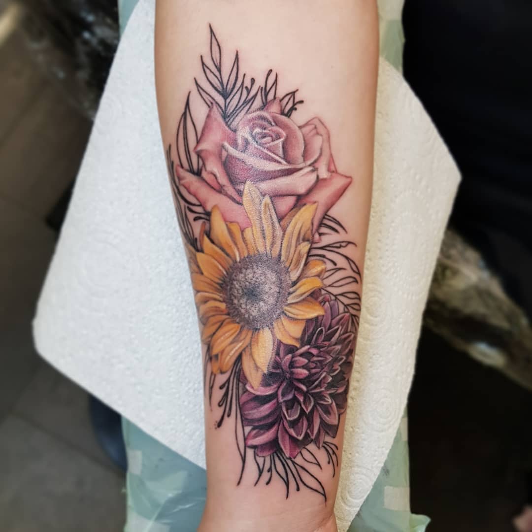 Forearm tattoo with detailed rose, sunflower, and dahlia in vibrant colors
