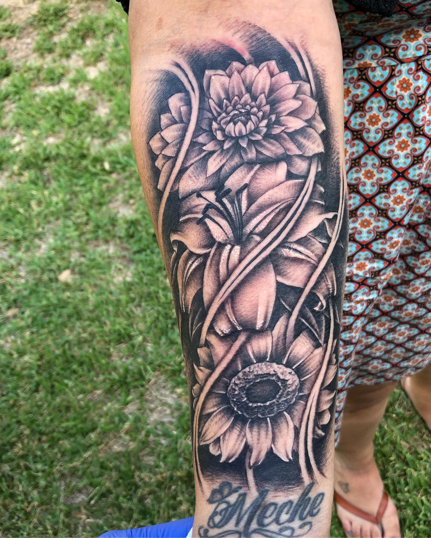 Black and grey forearm tattoo with detailed dahlia, lily, and sunflower flowers in a bold design