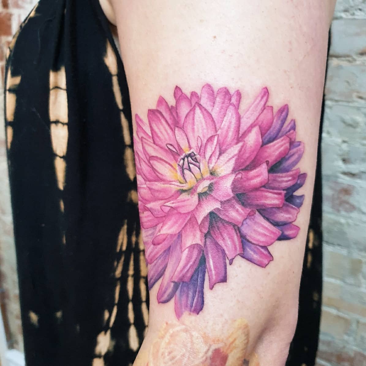 Vibrant pink dahlia flower tattoo with detailed shading and a yellow center on the upper arm