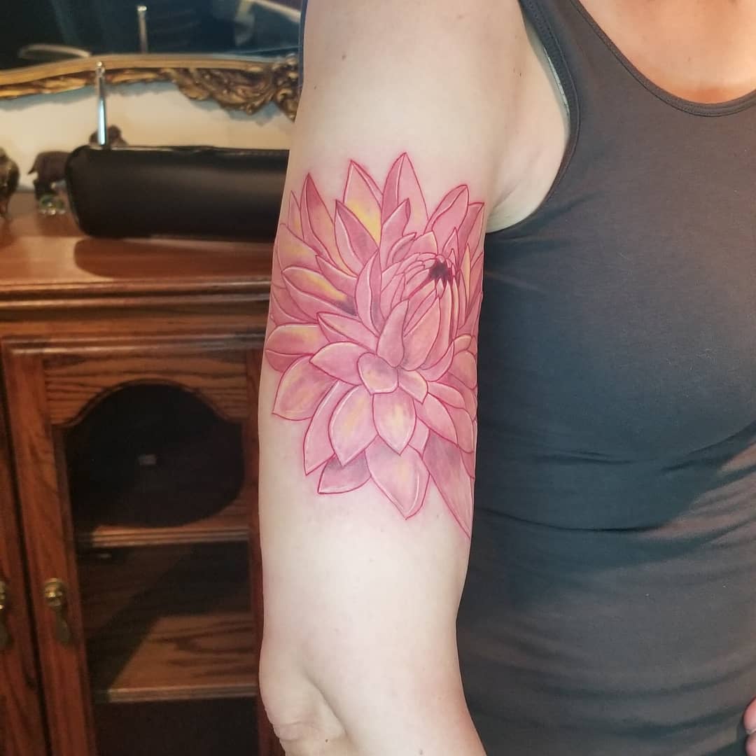 Colorful upper arm tattoo featuring a large pink dahlia with soft yellow accents on the petals