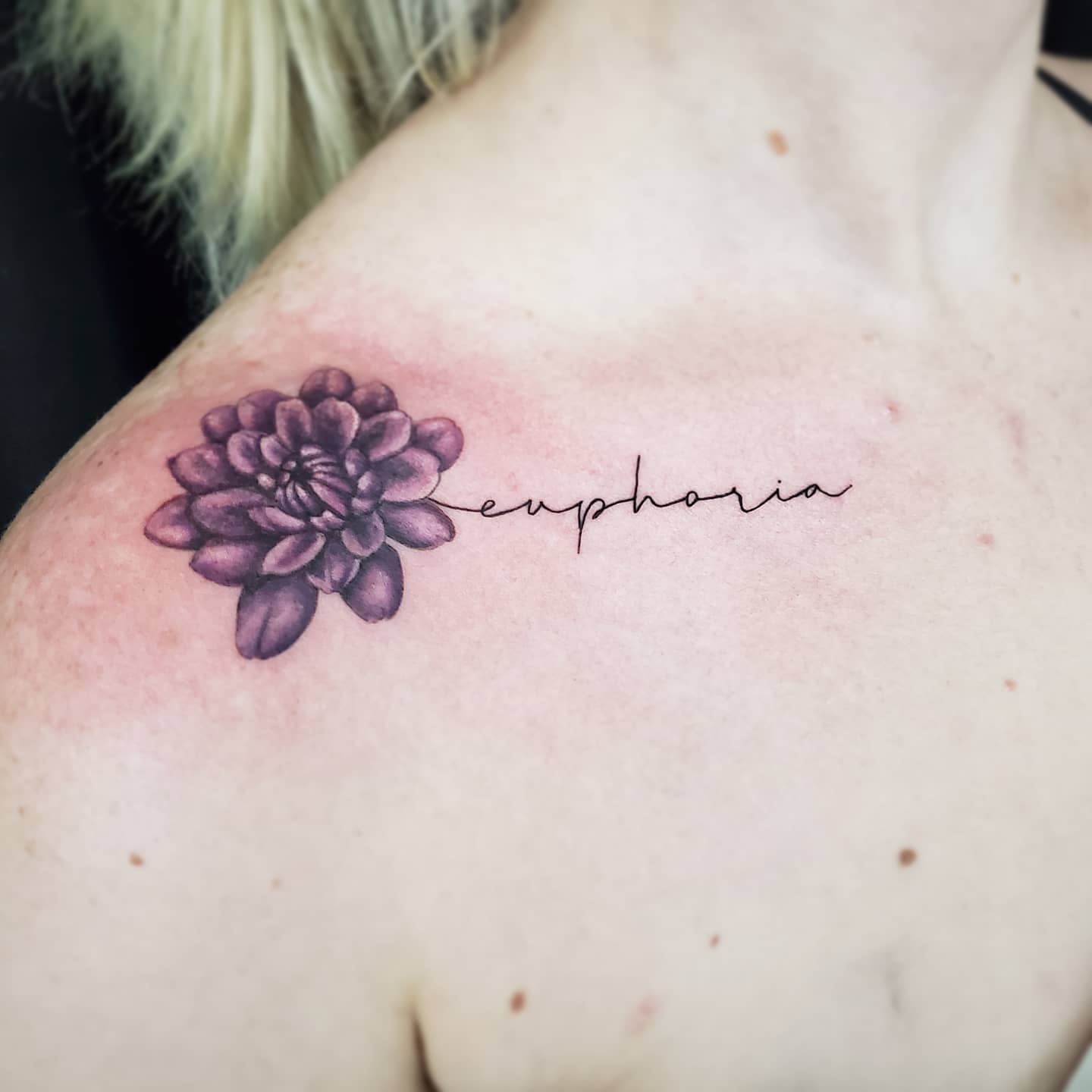 Small purple dahlia tattoo on the upper chest with the word "euphoria" in cursive script