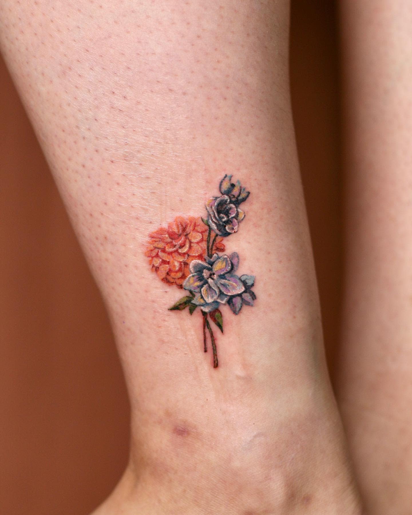 Small and delicate tattoo on the lower leg featuring an orange dahlia and two smaller flowers
