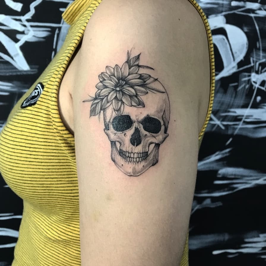 Black and grey upper arm tattoo featuring a realistic skull with a dahlia flower on top