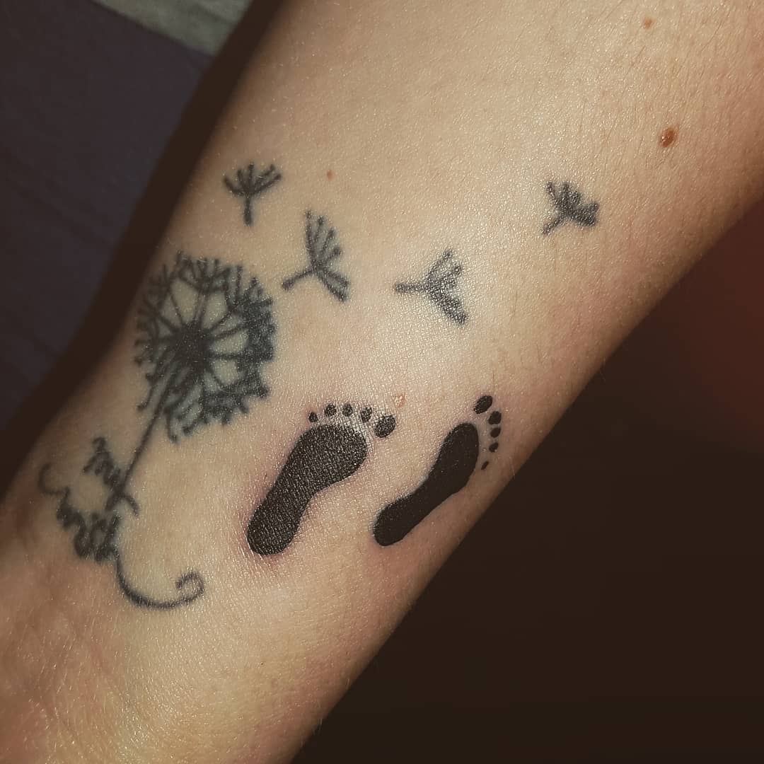 Tattoo of a dandelion with seeds blowing away and two baby footprints on an arm