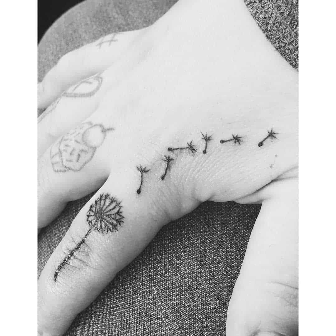 Black and white tattoo of a dandelion blowing in the wind on the back of a hand