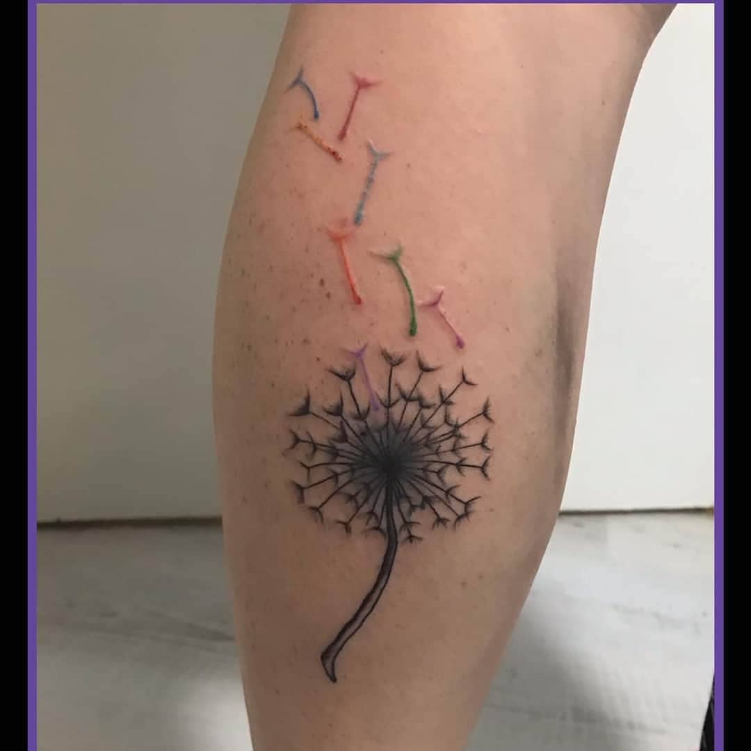 Tattoo of a dandelion with colorful seeds blowing away inked on the back of an arm