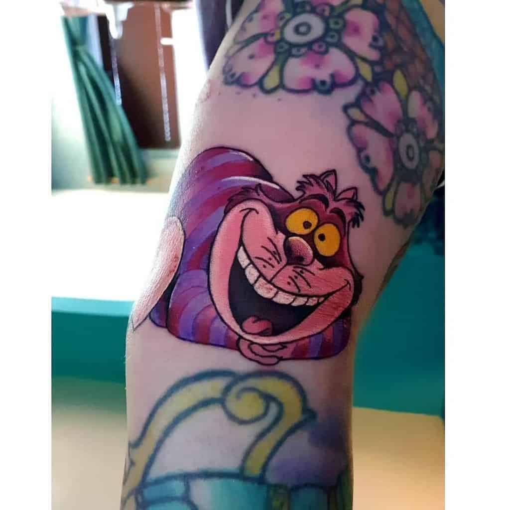 A colorful tattoo of the classic Disney Cheshire Cat, featuring its wide grin and iconic pink and purple stripes
