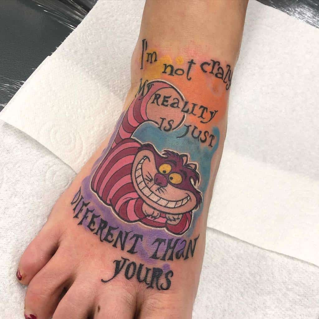 A colorful Cheshire Cat tattoo on the foot with the quote, "I'm not crazy, my reality is just different than yours"