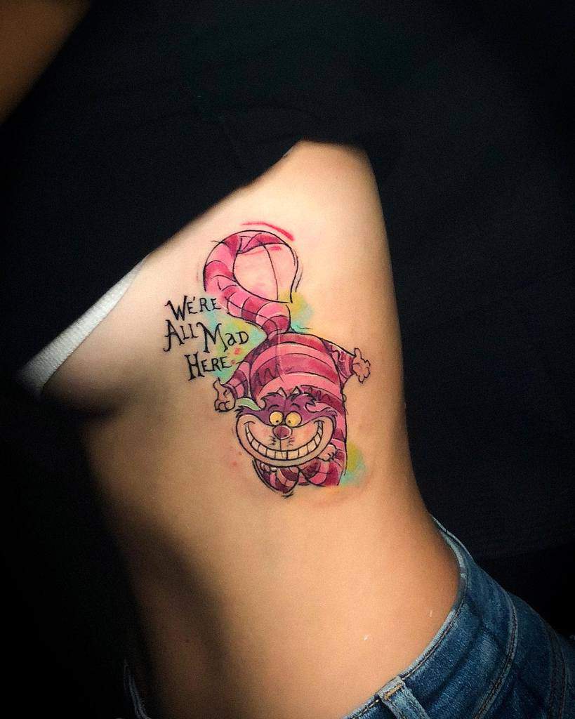 A colorful Cheshire Cat tattoo on the side with the quote "We're all mad here," featuring its iconic grin and pink stripes