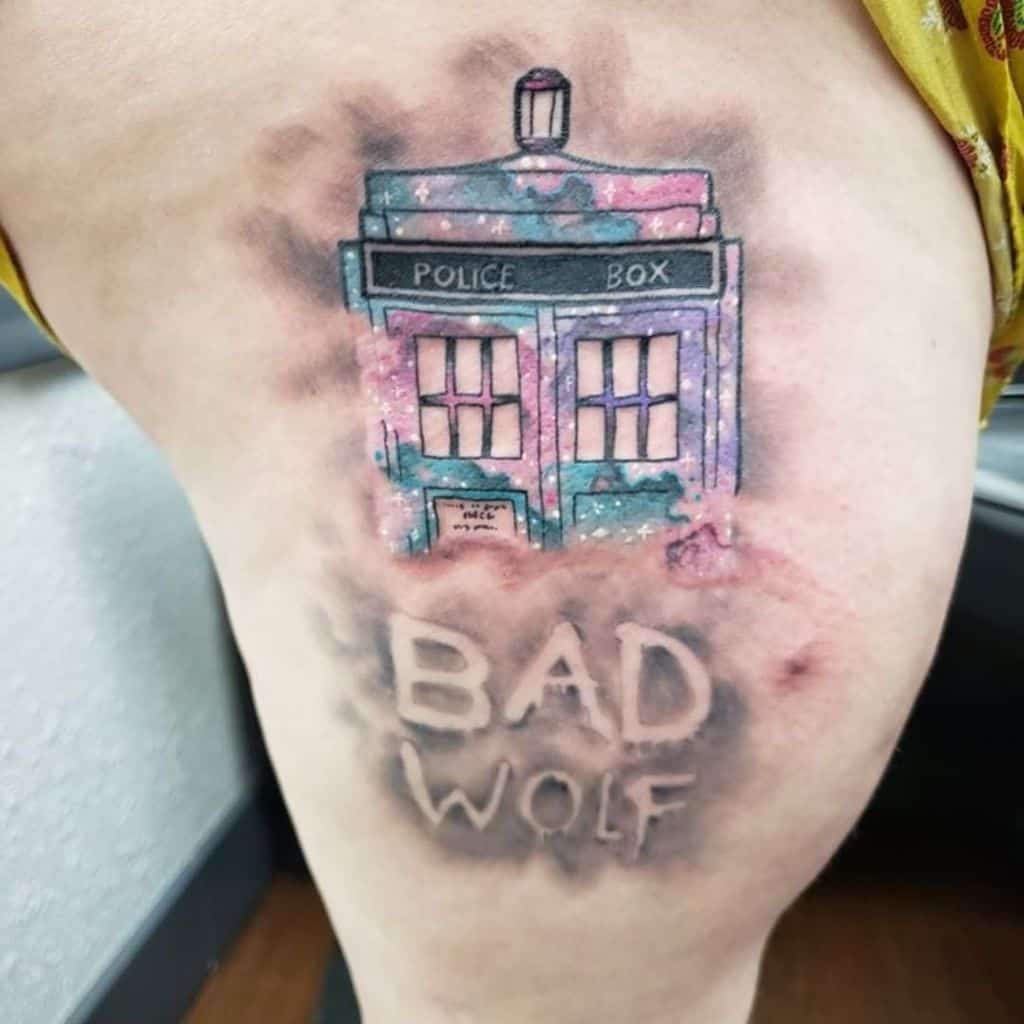 Watercolor tattoo on a thigh featuring the TARDIS from 'Doctor Who' amidst a pink and purple cosmic mist, with 'BAD WOLF' subtly inscribed below in a cloud-like formation