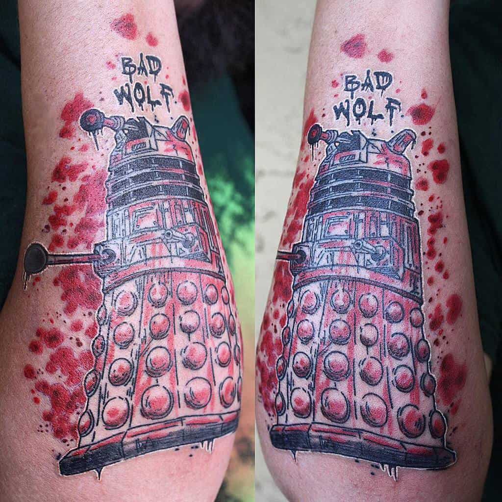 Detailed tattoo on an arm depicting a Dalek from 'Doctor Who' in red and black ink, splattered with red to resemble blood, with 'BAD WOLF' graffitied above it