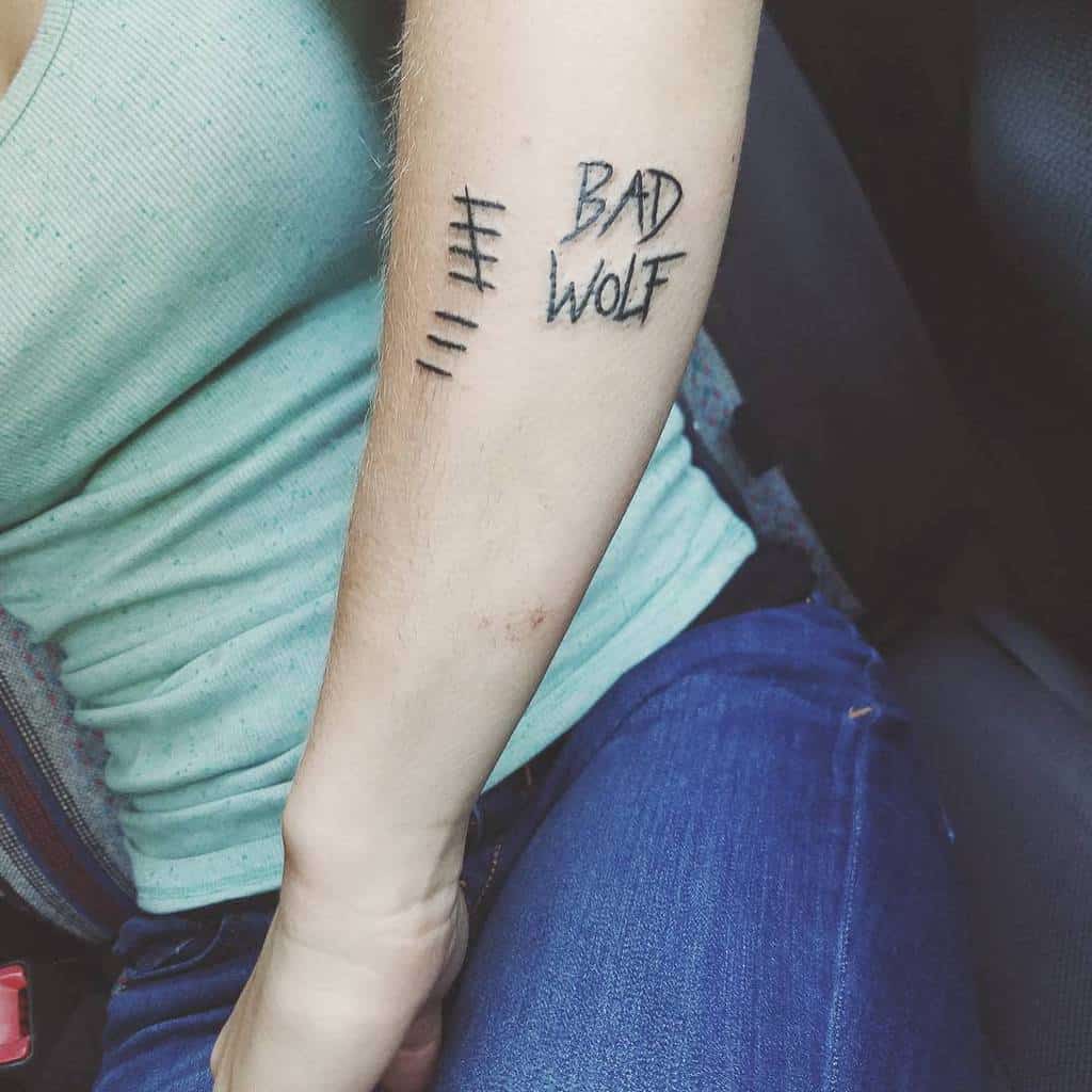 Minimalist black ink tattoo on an arm featuring the phrase 'BAD WOLF' with tally marks, symbolizing a recurring theme from 'Doctor Who'