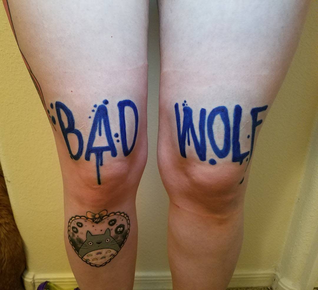 Pair of leg tattoos displaying 'BAD WOLF' in bold blue dripping letters on each calf, with a cute heart-shaped Totoro tattoo positioned below on one leg