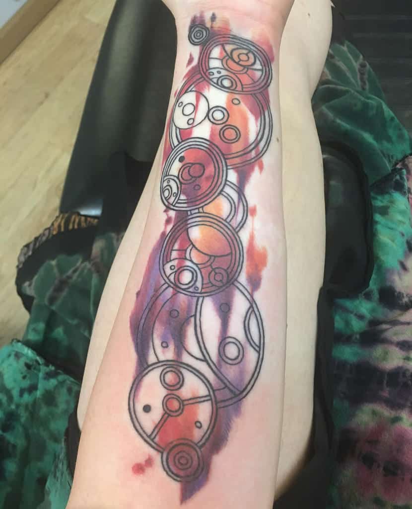 Vibrant tattoo featuring colorful Gallifreyan script from 'Doctor Who', depicted as a series of interlinked circles and intricate designs, set against a dynamic watercolor background in shades of red and purple