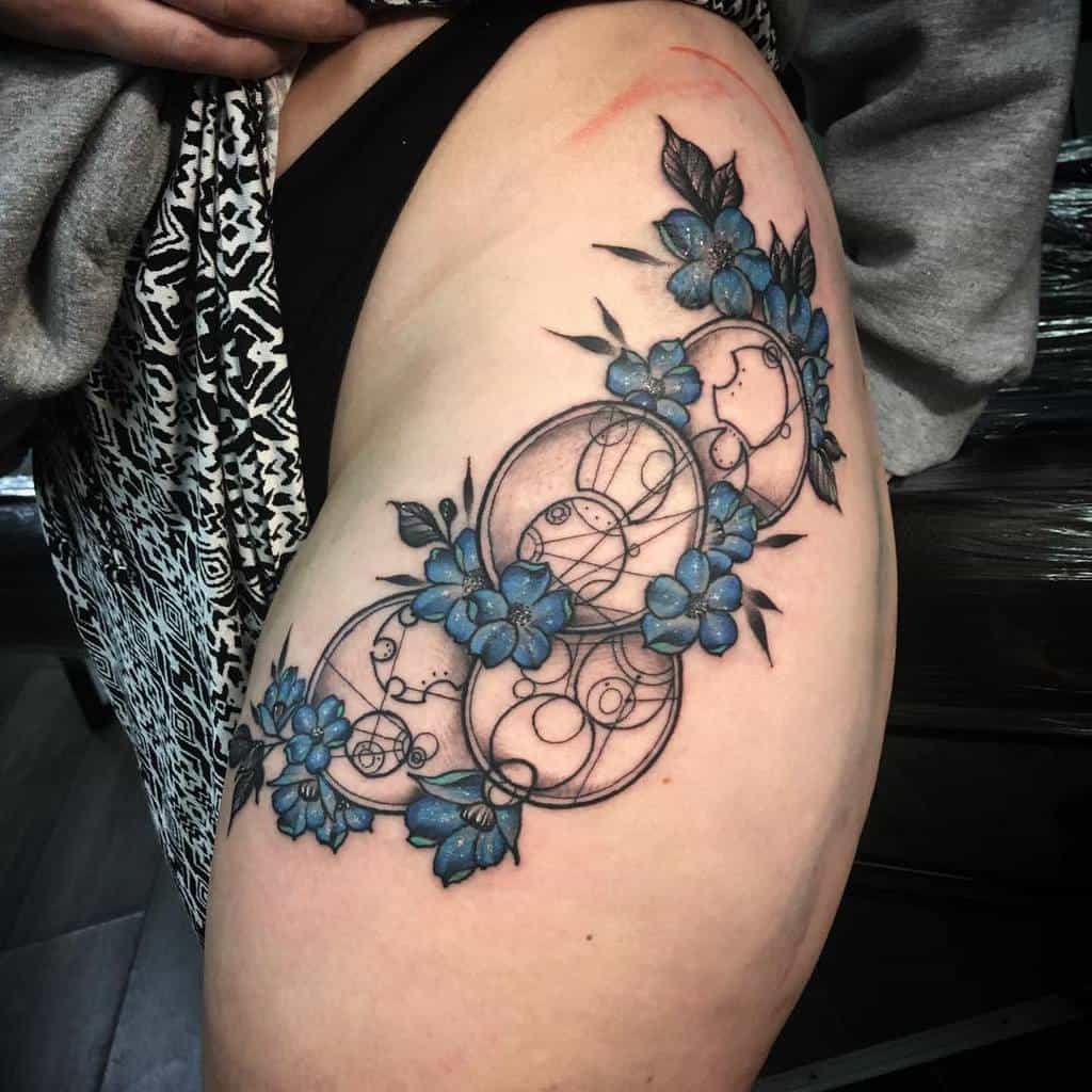 Tattoo of Gallifreyan script from 'Doctor Who' intertwined with a garland of blue flowers, blending intricate black circles with vibrant floral accents