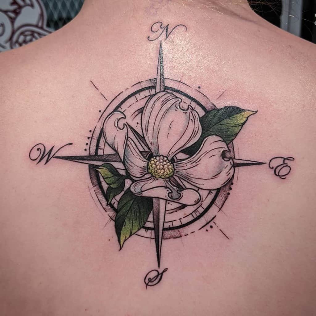 A back tattoo of a compass rose with a vibrant dogwood flower at the center, featuring green leaves and cardinal directions