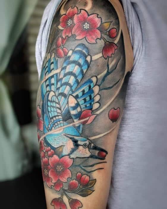 Vibrant tattoo featuring a blue jay in flight with pink dogwood flowers and branches, covering the upper arm