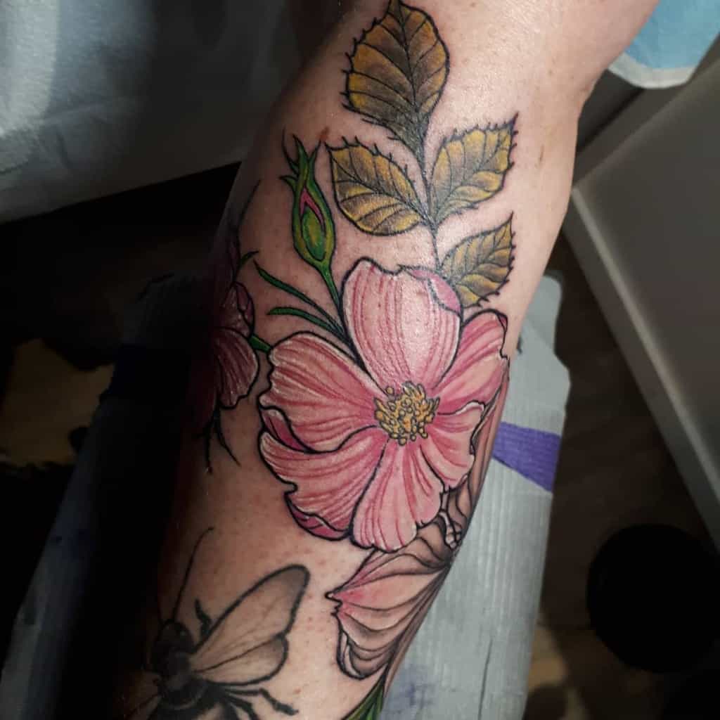 A tattoo of pink dogwood flowers, vibrant green leaves, and a bee adorns an arm