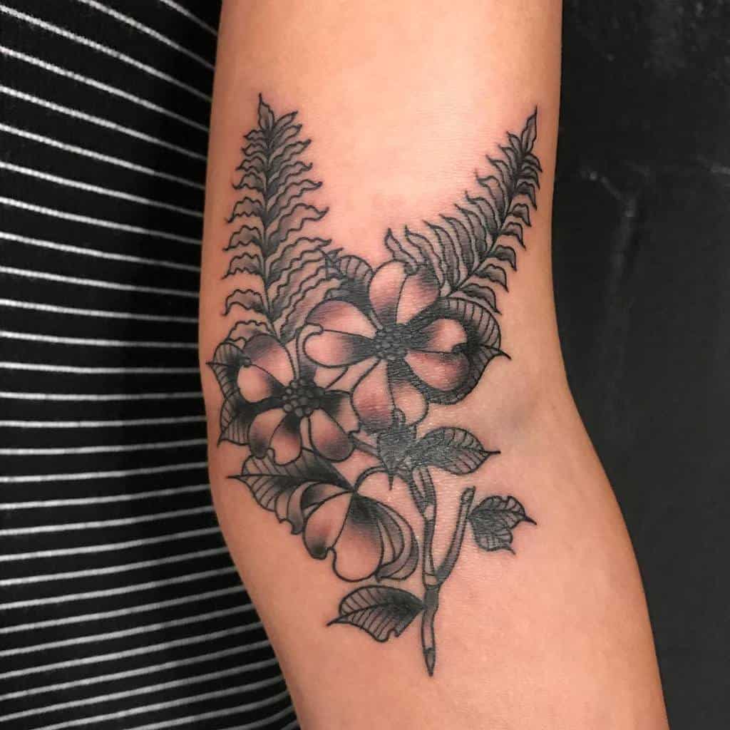 A black dogwood and fern tattoo elegantly wraps around a person's arm, creating a stunning floral design