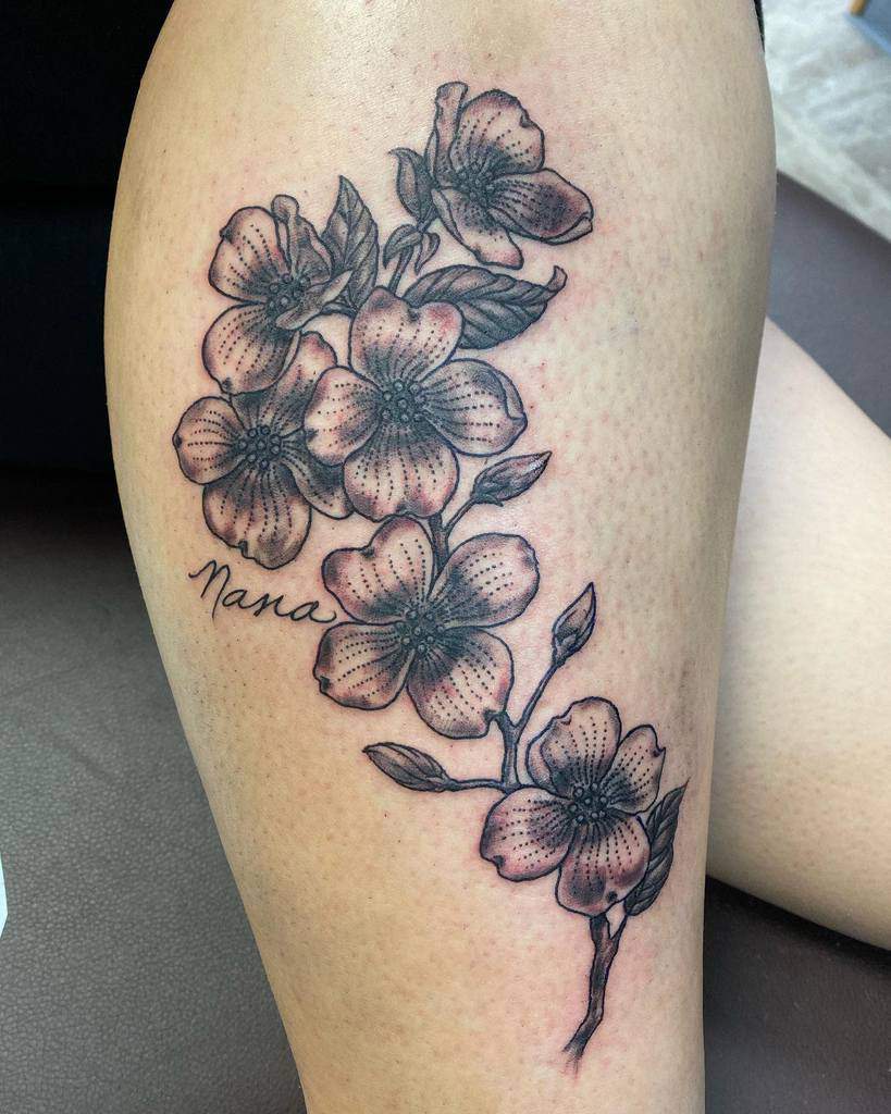 A tattoo featuring delicate dogwood flowers and leaves adorns a leg, with the heartfelt inscription "Nana" nestled beside it
