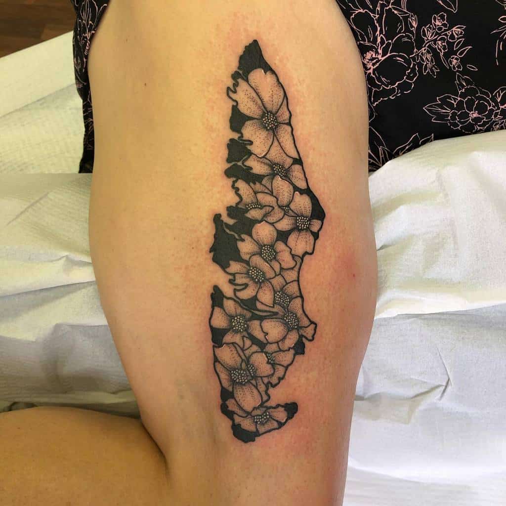 Black ink tattoo of dogwood flowers arranged in the shape of a state or geographic outline, placed on the upper leg