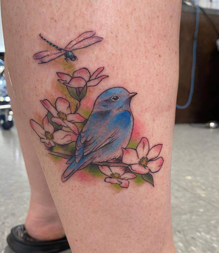 A tattoo of a blue bird, pink dogwood flowers, and a dragonfly adorns a leg