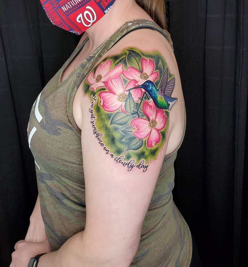 A colorful hummingbird and dogwood flowers tattoo adorn a woman's upper arm with text: "I've got sunshine on a cloudy day"
