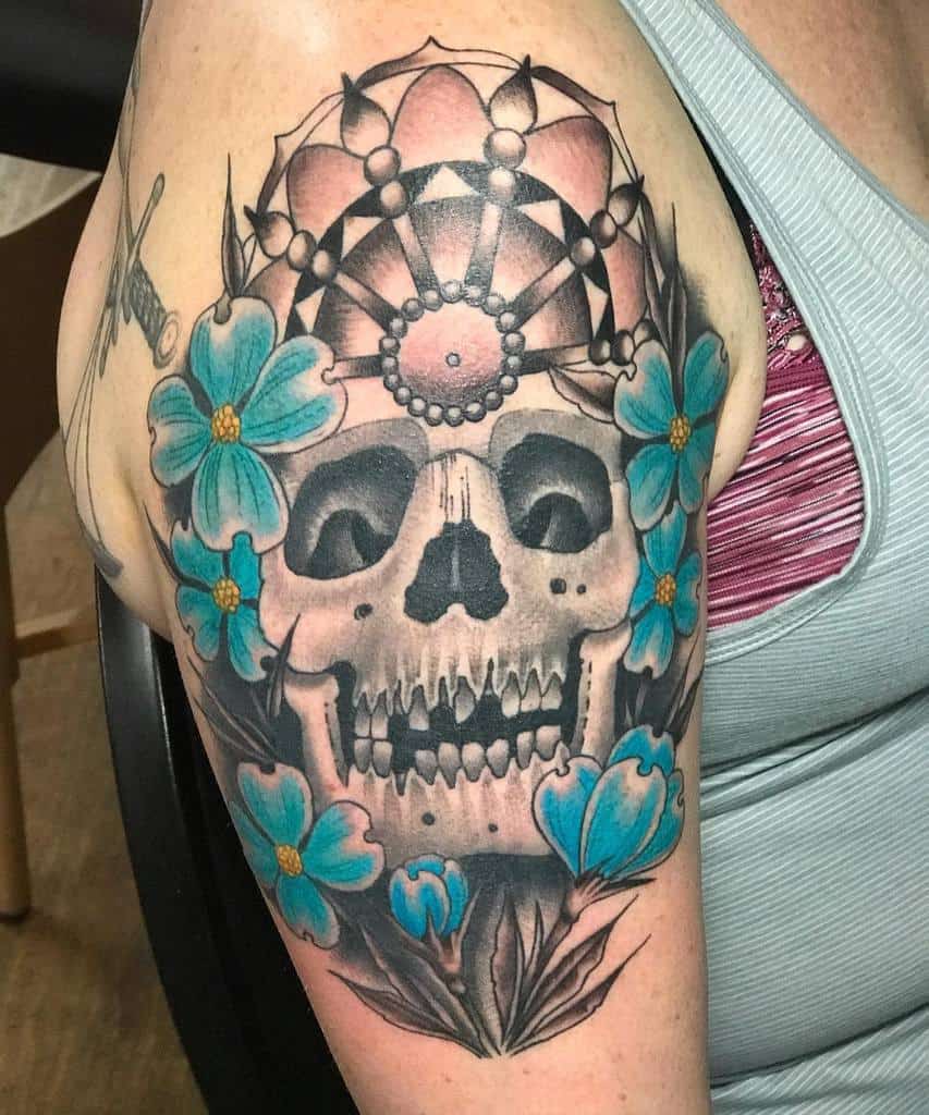 Tattoo of a skull adorned with a crown, surrounded by blue dogwood flowers on an upper arm