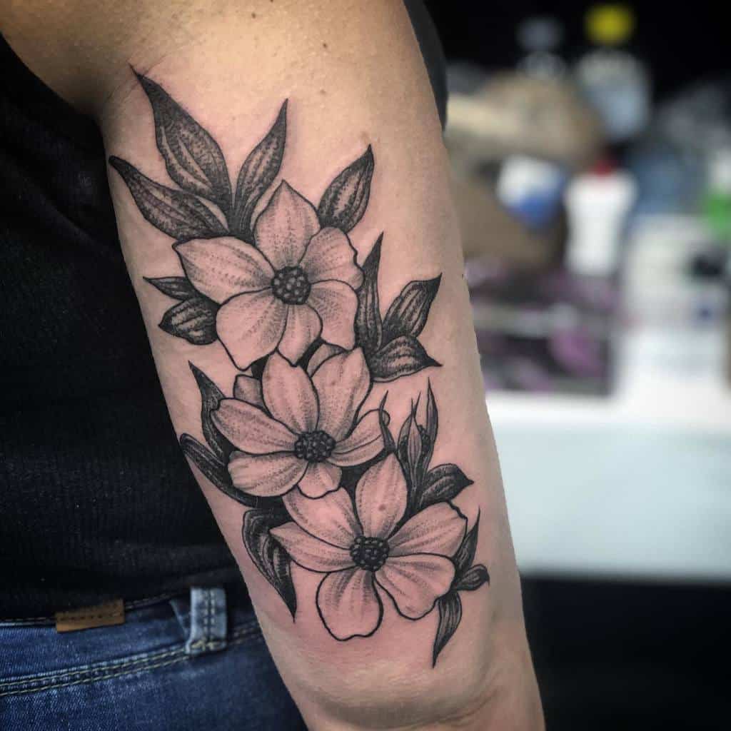 A stunning arm tattoo featuring black and gray dogwood flowers and intricate leaves graces the skin