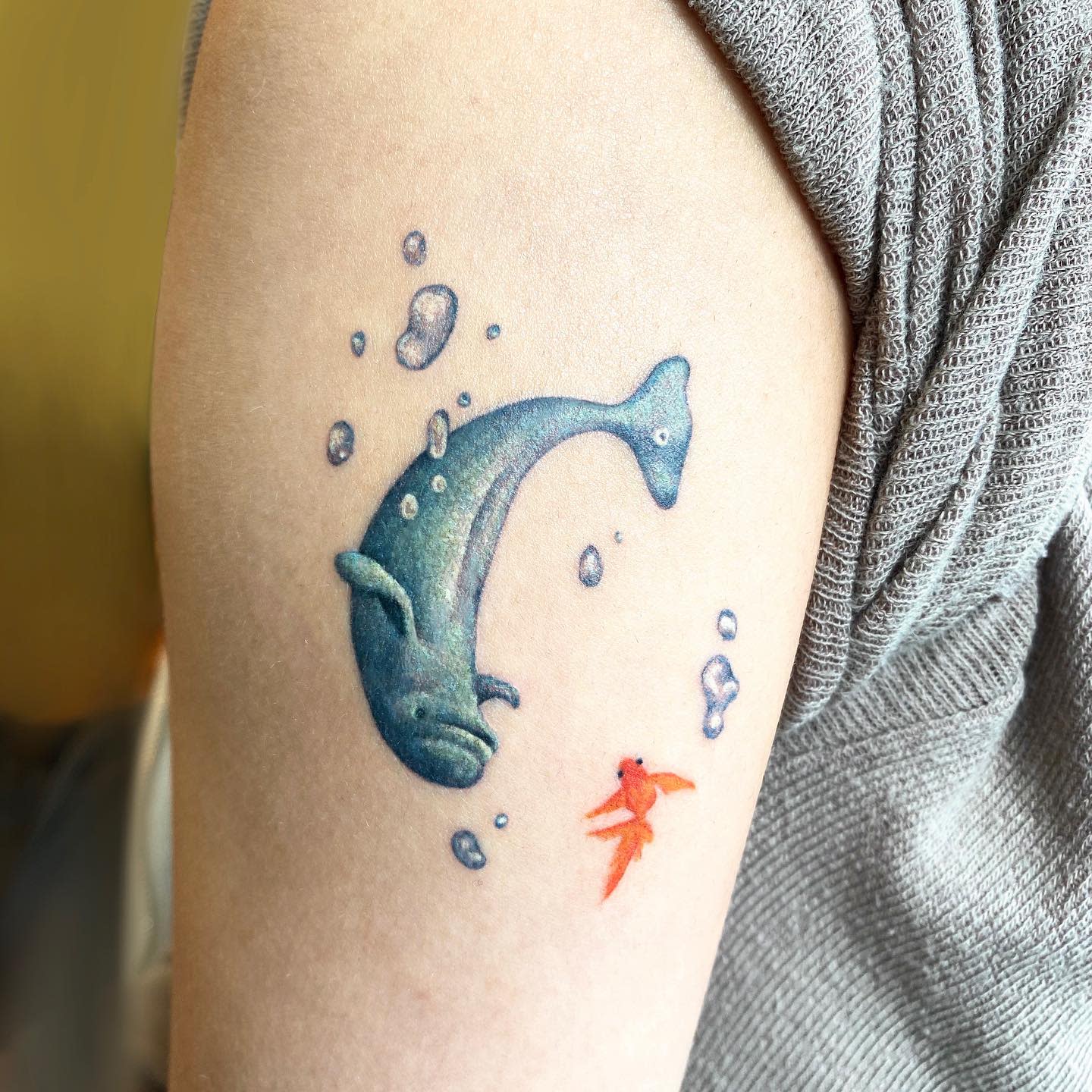 Tattoo of a small blue whale and an orange fish with bubbles on an upper arm