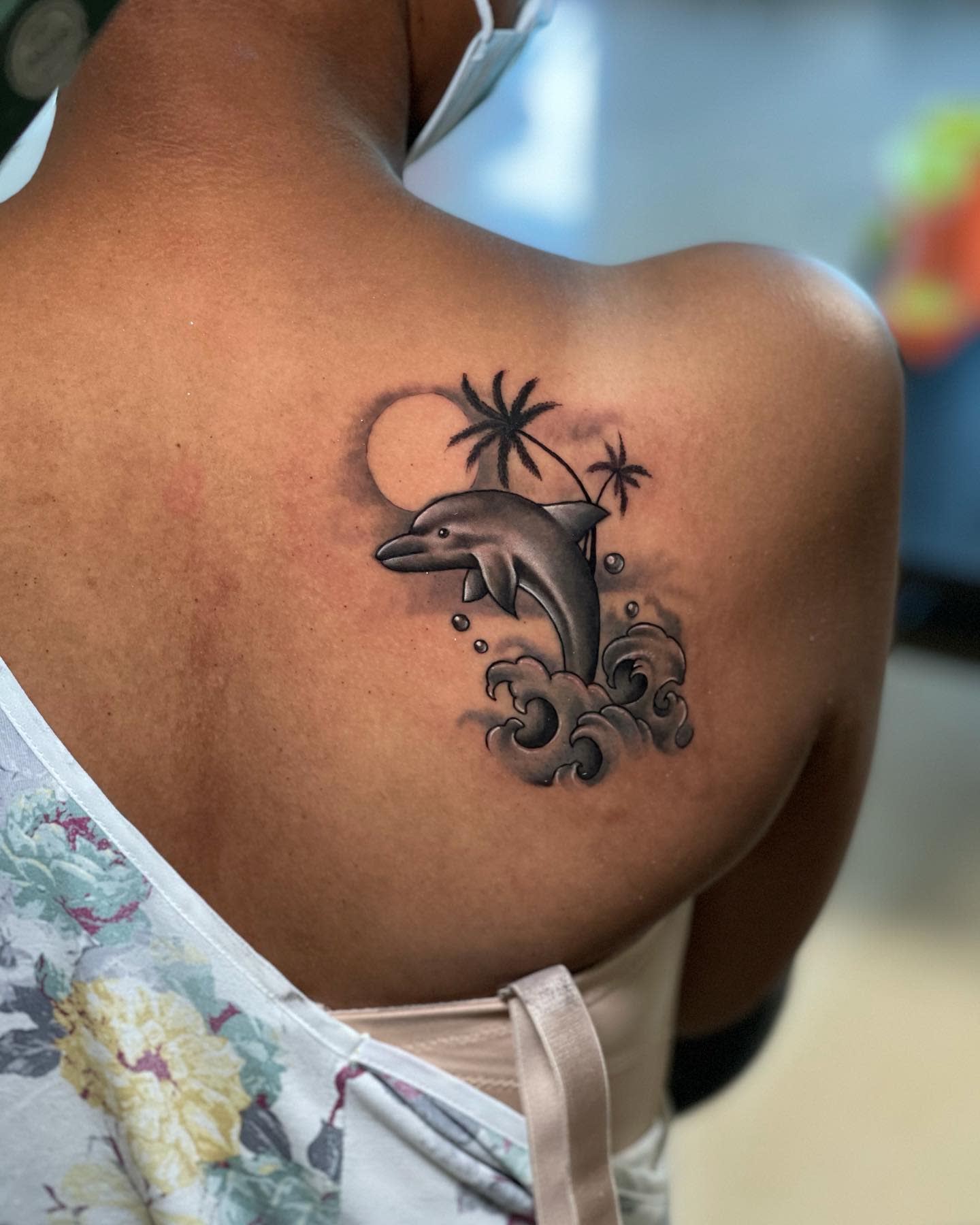 A tattoo of a dolphin, waves, and palm trees on a woman's upper back and shoulder