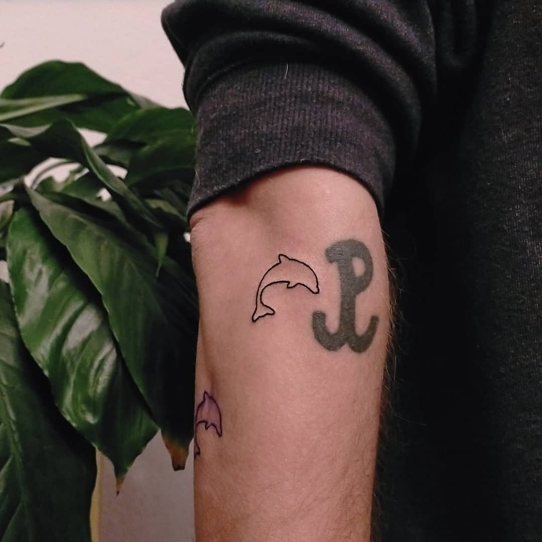 Arm featuring three tattoos: a large "P", a black dolphin, and a small purple dolphin